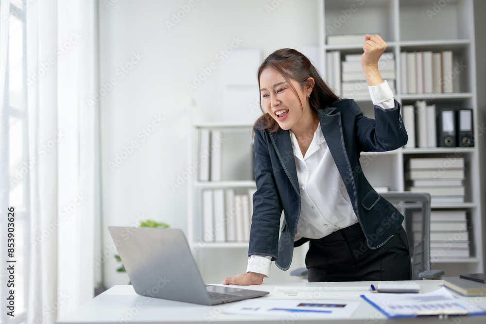 Excited businesswoman celebrating success at work while looking at laptop in modern office setting with documents on desk. Excited businesswoman celebrating success at work while looking at laptop in modern office setting with documents on desk.
