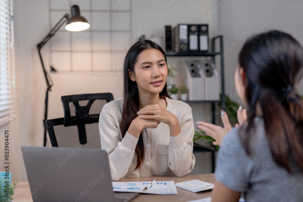 Two women having a professional conversation in a modern office setting, one taking notes on a laptop while the other listens attentively. Two women having a professional conversation in a modern office setting, one taking notes on a laptop while the other listens attentively.
