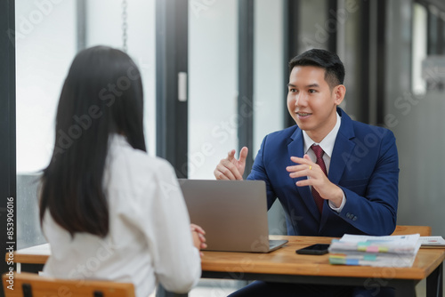 Two professionals in a business meeting discussing work at a table, engaging in conversation with a laptop and documents present.