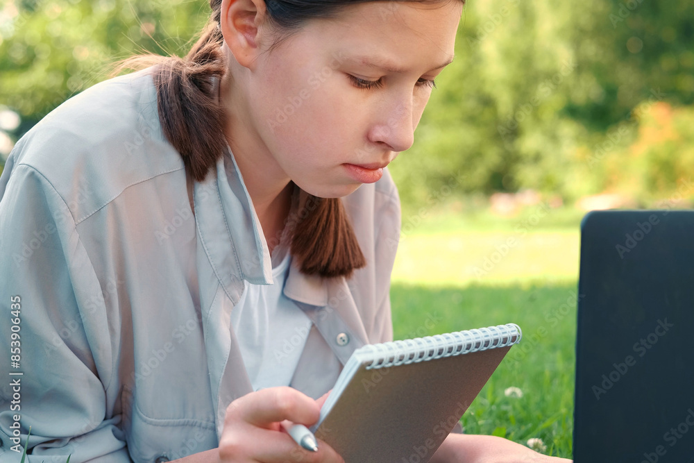 Fototapeta premium Teenage schoolgirl studying reading her books, tablet and notebook, sitting outdoors. Back to school. Student girl lying on the green grass using laptop in the college yard or park. Distance learning.