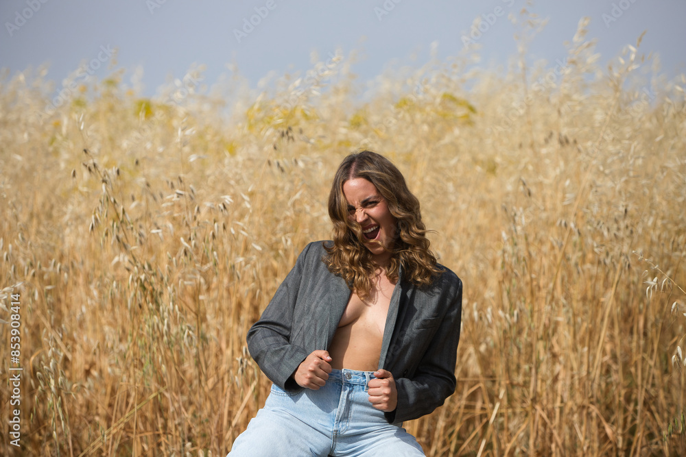 Young, pretty blonde woman is dressed in grey jacket, jeans, boots sitting on a stool in a field planted with oats. The woman is shouting with rage and anger. Young, pretty blonde woman is dressed in grey jacket, jeans, boots sitting on a stool in a field planted with oats. The woman is shouting with rage and anger.