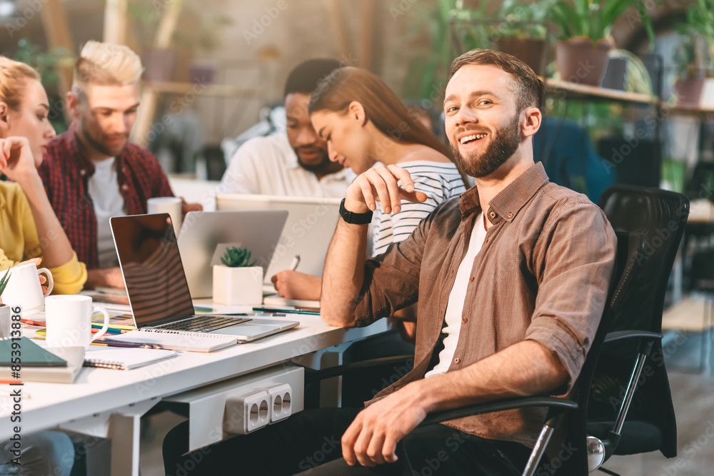 A smiling man sits at a desk in a modern office while his colleagues engage in a collaborative work session around him. A smiling man sits at a desk in a modern office while his colleagues engage in a collaborative work session around him.
