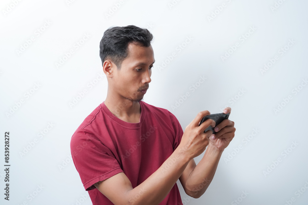 Young Asian man posing holding a cellphone with both hands, wearing a red t-shirt isolated on a white background. Shot in studio. Gaming concept. Young Asian man posing holding a cellphone with both hands, wearing a red t-shirt isolated on a white background. Shot in studio. Gaming concept.