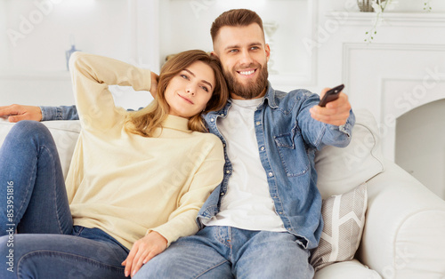 A cheerful couple sits on a comfortable sofa, enjoying their time watching television together in a warm, well-decorated living room. © Prostock-studio