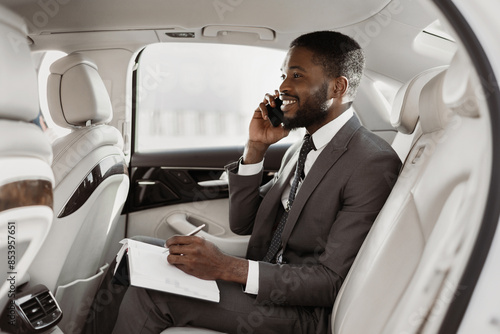 Black businessman is sitting in the backseat of a car, taking notes on a notepad while talking on a phone. He is wearing a suit and tie, and he has a serious expression on his face. © Prostock-studio