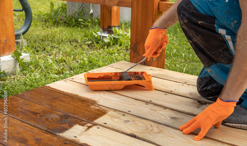 Wallpaper Mural A worker applies a brown stain to a wooden deck with an orange roller tray and brush. The stain is visible on the wood, and the worker is wearing gloves for protection. Torontodigital.ca