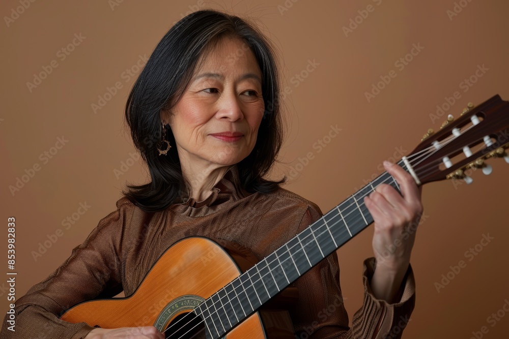 Portrait of a tender asian woman in her 50s playing the guitar while standing against pastel brown background Portrait of a tender asian woman in her 50s playing the guitar while standing against pastel brown background