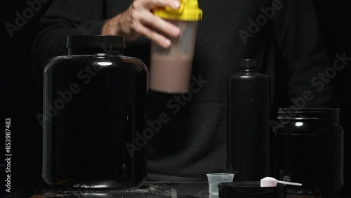 A Man Shakes Chocolate Protein In A Shaker And Tastes It. Next To Him Are Black Cans With Vitamins And Supplements For Athletes. photo