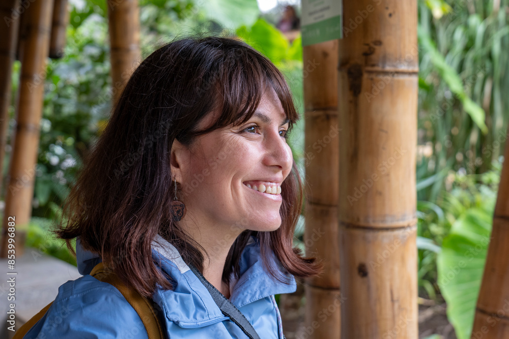 A Young Brown Woman in a Blue Coat with a Camera Strap Around her Neck Smiles and Stares at the Sky on A Rainy Day with Bamboo Poles in the Background at a Telephoto Angle