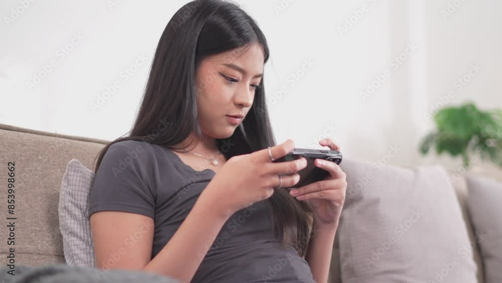 A young Asian woman is sitting on a couch, playing a game on her smartphone with an excited expression. She raises her fist in a gesture of triumph A young Asian woman is sitting on a couch, playing a game on her smartphone with an excited expression. She raises her fist in a gesture of triumph