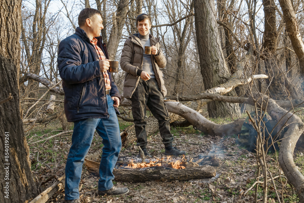 Two men make a campfire for cooking in the forest, father and son sitting on a log while hiking and outdoor activities, early spring landscape Two men make a campfire for cooking in the forest, father and son sitting on a log while hiking and outdoor activities, early spring landscape