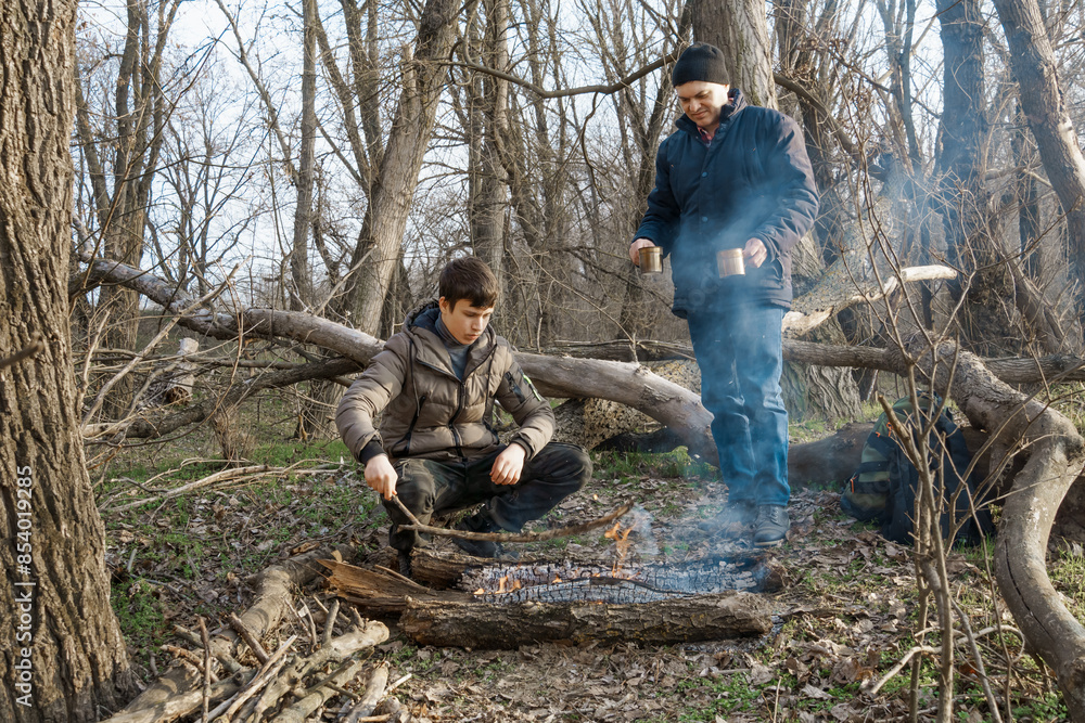 Two men make a campfire for cooking in the forest, father and son sitting on a log while hiking and outdoor activities, early spring landscape Two men make a campfire for cooking in the forest, father and son sitting on a log while hiking and outdoor activities, early spring landscape