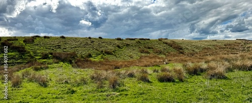 Rolling green hills beneath a dramatic, cloud-filled sky dominate the landscape. Various shades of lush grass weave a verdant tapestry across the moors of Sutton-in-Craven, UK