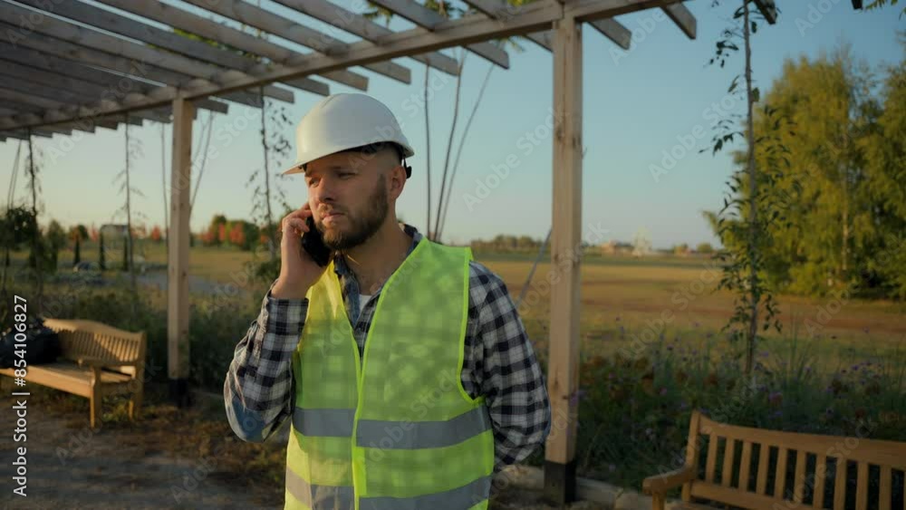 Serious Caucasian male worker man specialist builder guy engineer in hardhat helmet talking mobile phone call answering smartphone calling device speaking discuss strategy project ecology park plan Serious Caucasian male worker man specialist builder guy engineer in hardhat helmet talking mobile phone call answering smartphone calling device speaking discuss strategy project ecology park plan