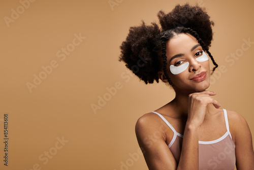 A young woman with dark curly hair and eye patches smiles confidently against a warm beige backdrop.