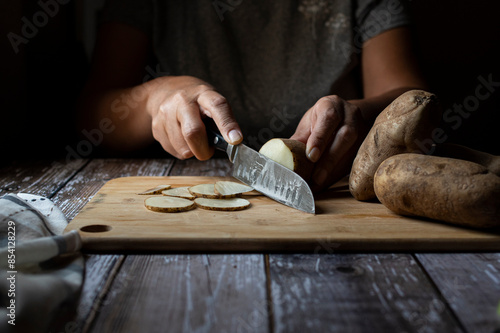 Concept of taking or grabbing food with hands, feminine Mexican hands. Cutting potatoes into slices on a wooden cutting board. horizontal dark mood close up image front view