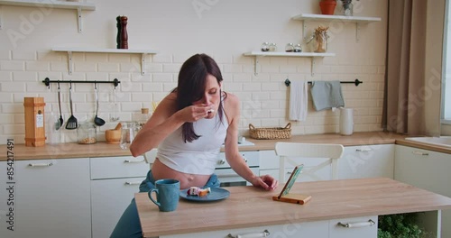 Hungry pregnant woman eat dessert at kitchen, pregnancy food cravings. Pregnancy enjoy sweet cake, sit on chair with belly