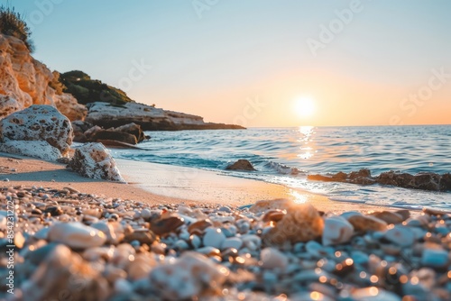 The sun sets over a calm sea, casting a warm glow on the pebbly shore of a tranquil beach