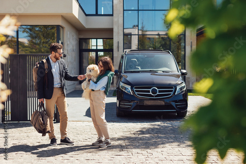 Young stylish couple hold dog in hands and walks from their luxury suburb house