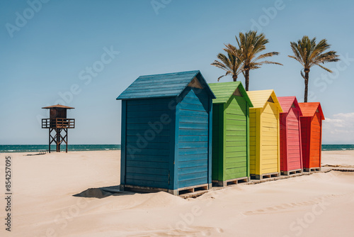 A row of brightly colored beach huts facing the sea, with palm trees and a lifeguard tower in the background. Sant Antoni beach, Cullera, Spain