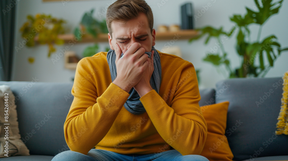 Young man sitting on the couch at home and coughing. Suffers from an attack of asthma, allergies. He holds his chest, covers his mouth with his hand Young man sitting on the couch at home and coughing. Suffers from an attack of asthma, allergies. He holds his chest, covers his mouth with his hand