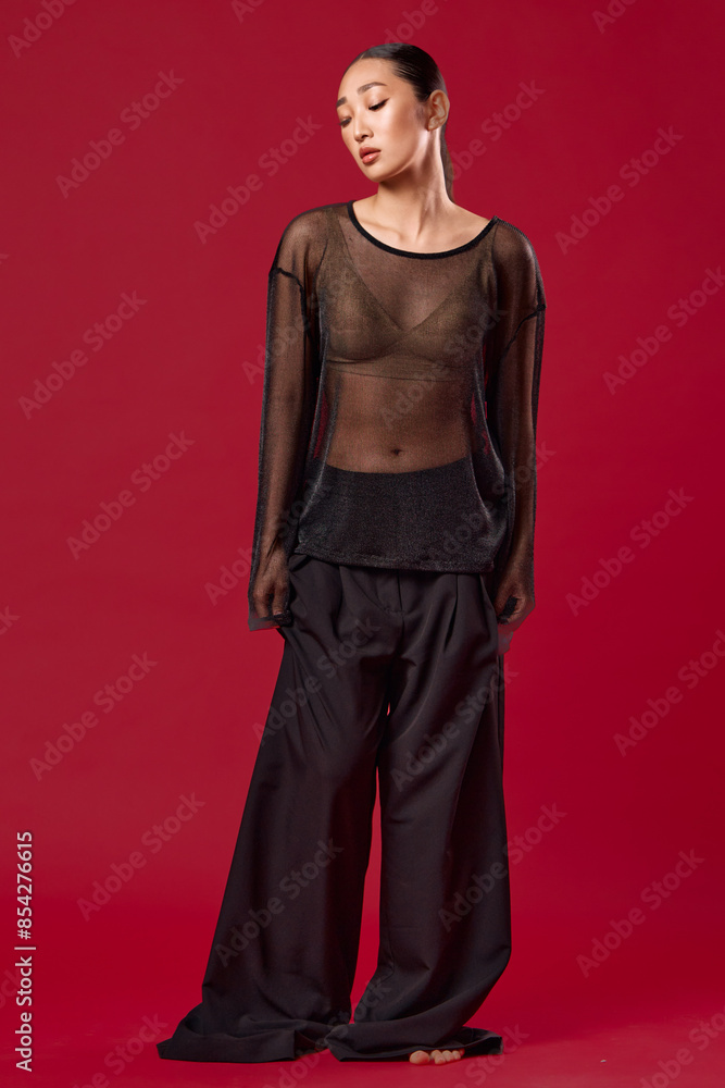 Fashion asian woman in sheer top and widelegged pants poses with hands on hips against red background Fashion asian woman in sheer top and widelegged pants poses with hands on hips against red background