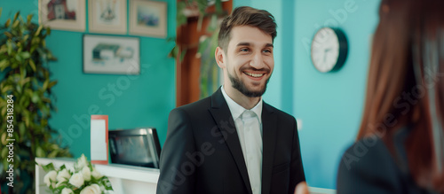 White male insurance company employee in stylish casual attire standing in front of a turquoise painted wall, warmly welcoming a client, demonstrating modern professional hospitality and customer ser