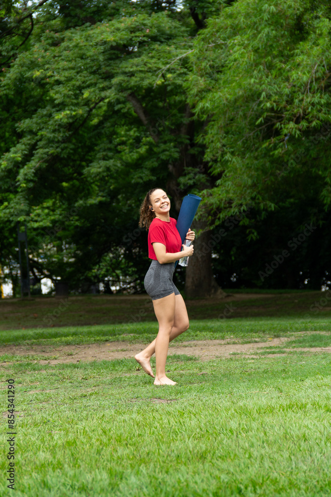 Smiling pretty happy woman holding yoga mat and water bottle barefoot in a public park outdoors. Yoga session and meditation concept Smiling pretty happy woman holding yoga mat and water bottle barefoot in a public park outdoors. Yoga session and meditation concept