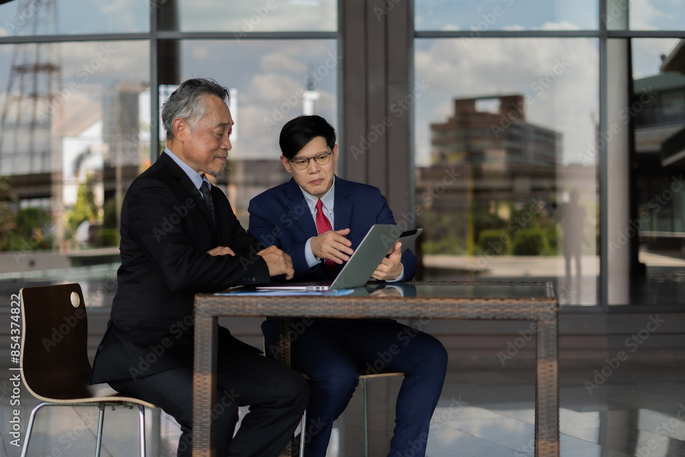 Two men in suits are looking at a laptop and tablet. They are discussing something important. Scene is serious and focused Two men in suits are looking at a laptop and tablet. They are discussing something important. Scene is serious and focused