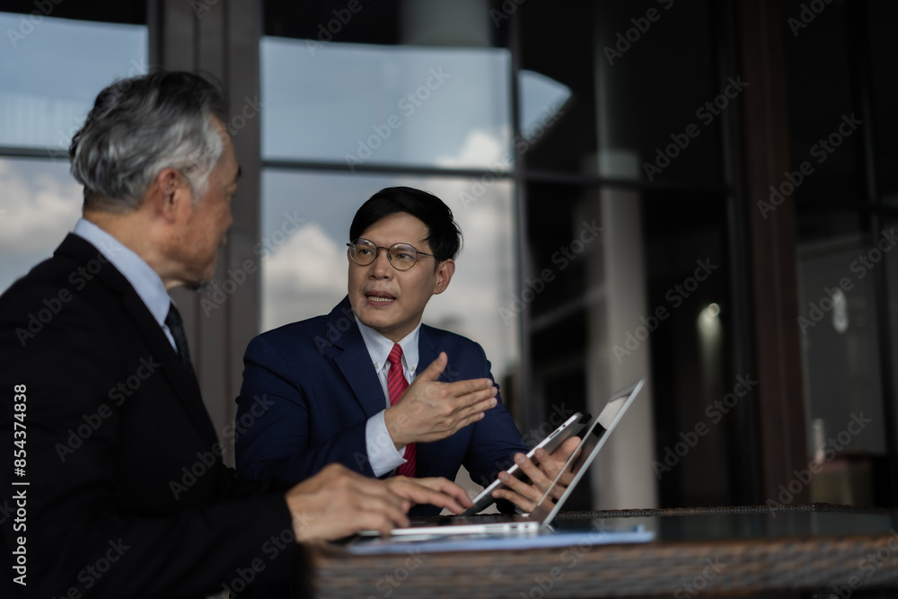 Two men in suits are looking at a laptop and tablet. They are discussing something important. Scene is serious and focused Two men in suits are looking at a laptop and tablet. They are discussing something important. Scene is serious and focused