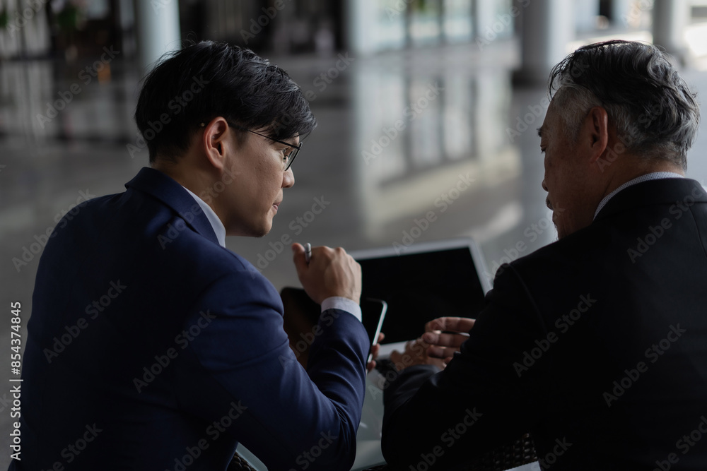 Two men in suits are looking at a laptop and tablet. They are discussing something important. Scene is serious and focused Two men in suits are looking at a laptop and tablet. They are discussing something important. Scene is serious and focused
