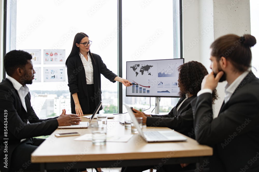 Business woman gives report to multiracial colleagues standing near graphs on monitor. Team meeting with analytics and performance charts. Professional setting with diverse team. Business woman gives report to multiracial colleagues standing near graphs on monitor. Team meeting with analytics and performance charts. Professional setting with diverse team.