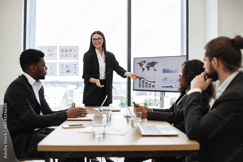 Fototapeta premium Business woman gives report to multiracial colleagues standing near graphs on monitor during office meeting. Professional business presentation with diverse team discussing data and strategies.