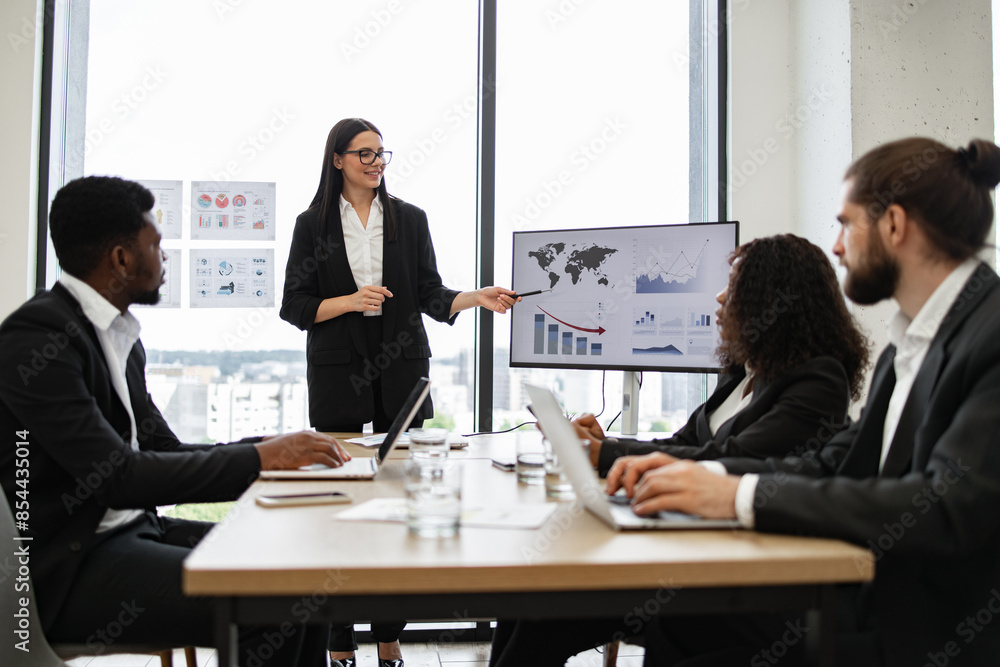 Business woman gives report to multiracial colleagues standing near graphs on monitor. Professional team discussing data analysis, growth charts, and performance metrics in modern office setting. Business woman gives report to multiracial colleagues standing near graphs on monitor. Professional team discussing data analysis, growth charts, and performance metrics in modern office setting.