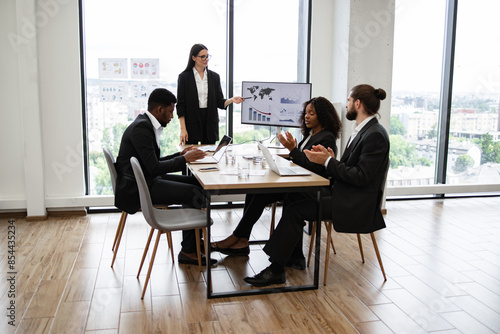 Caucasian business woman makes report to multiracial colleagues standing near monitor with graphs, charts. Diverse business people clap their hands. Modern office with windows showcasing city view.