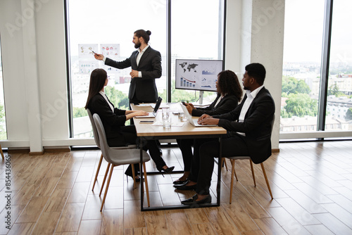 Young bearded businessman giving presentation to multiracial colleagues standing near graphs and charts on window glass. Modern office with diverse colleagues sitting at table with laptops.
