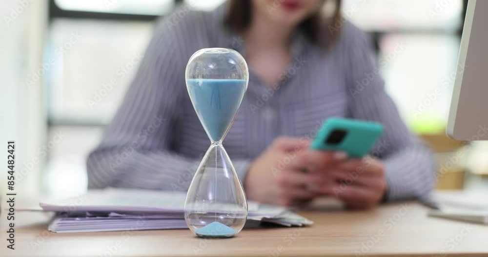 Woman uses mobile phone at desk with hourglass in office Woman uses mobile phone at desk with hourglass in office