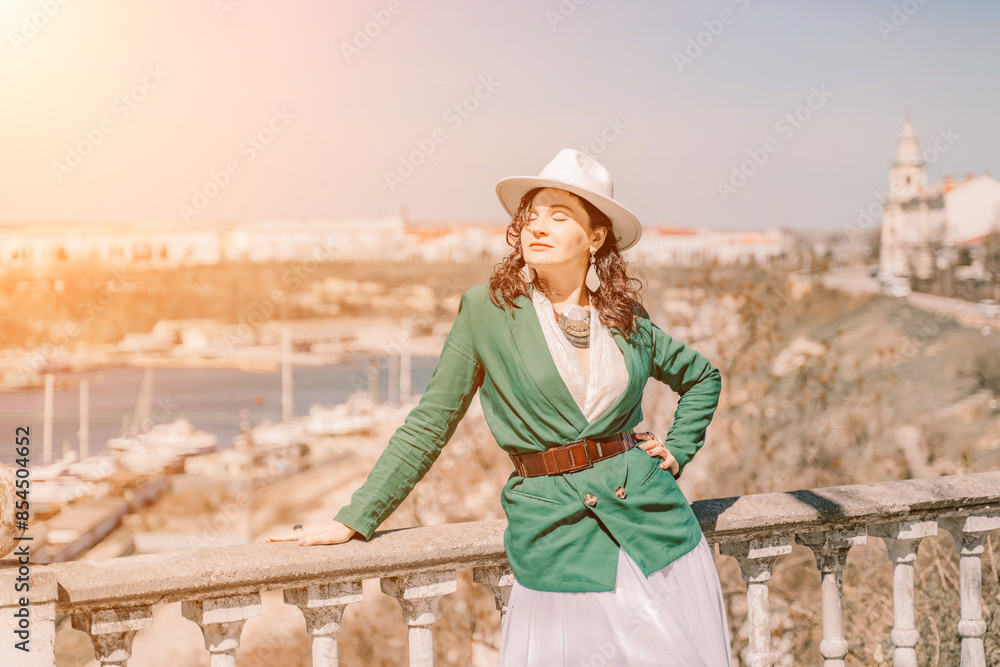 Obraz premium Woman walks around the city, lifestyle. Happy woman in a green jacket, white skirt and hat is sitting on a white fence with balusters overlooking the sea bay and the city.