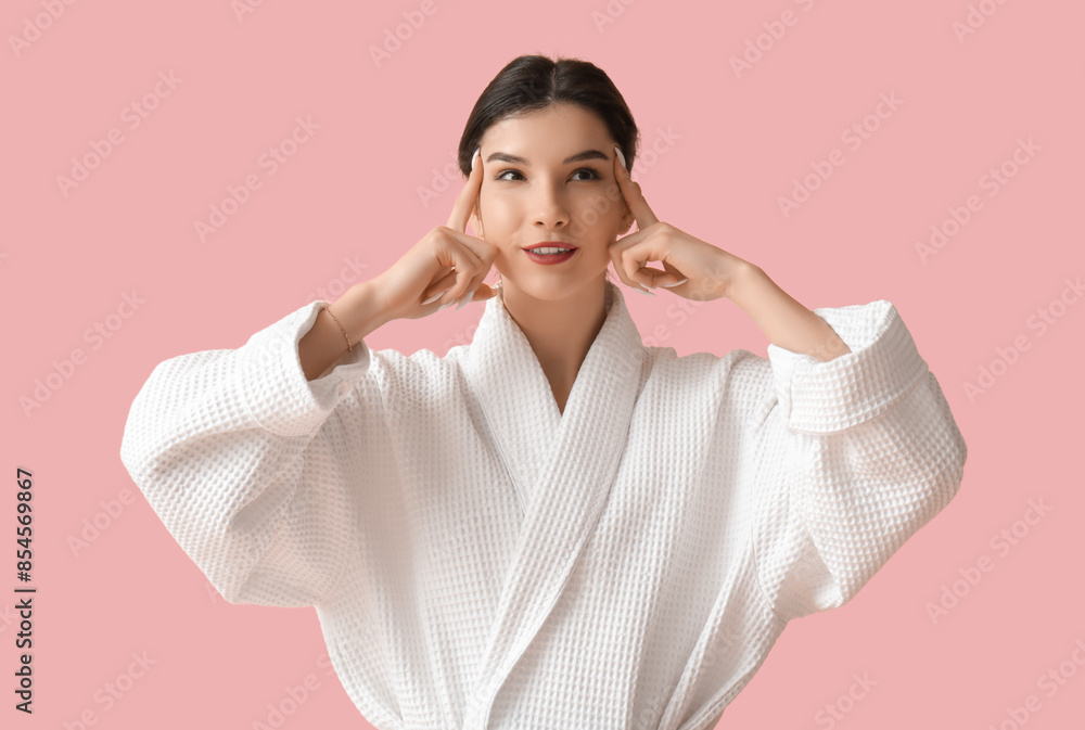 Beautiful young woman in bathrobe doing face building exercise on pink background Beautiful young woman in bathrobe doing face building exercise on pink background
