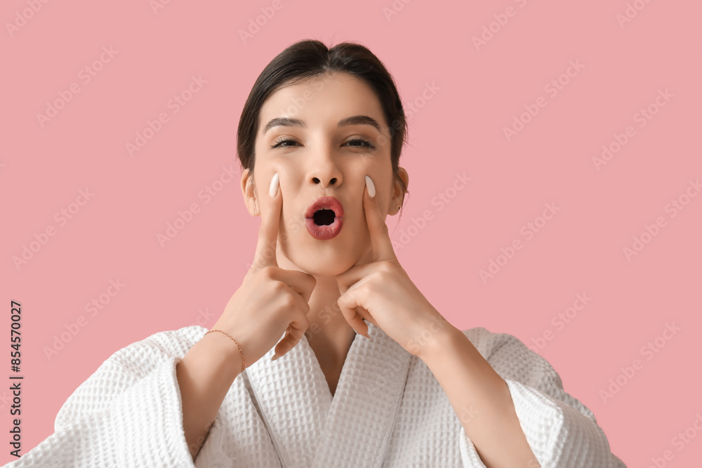 Beautiful young woman in bathrobe doing face building exercise on pink background Beautiful young woman in bathrobe doing face building exercise on pink background