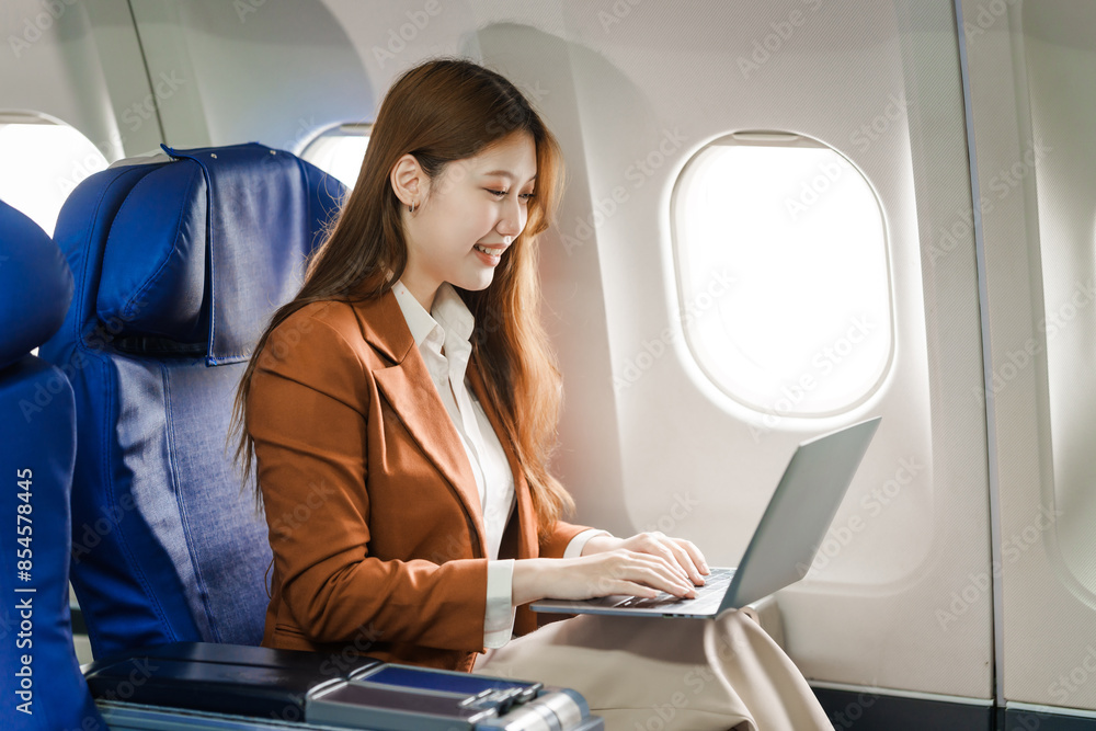 A young Asian businesswoman, dressed in a formal suit, sits at her airplane seat with a laptop and briefcase, utilizing in-flight Wi-Fi for a conference call. A young Asian businesswoman, dressed in a formal suit, sits at her airplane seat with a laptop and briefcase, utilizing in-flight Wi-Fi for a conference call.