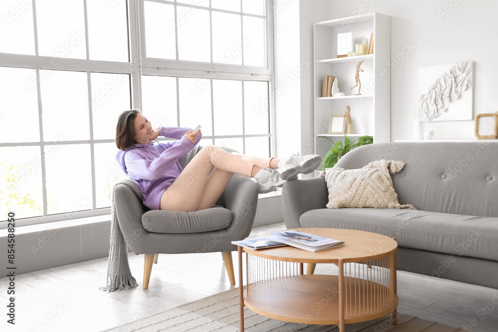 Smiling young woman with air conditioner remote control sitting on armchair in living room Smiling young woman with air conditioner remote control sitting on armchair in living room
