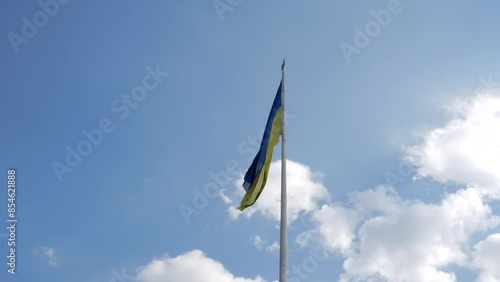 The flag of Ukraine flutters slowly on a flagpole with a coat of arms against the background of the sky and clouds