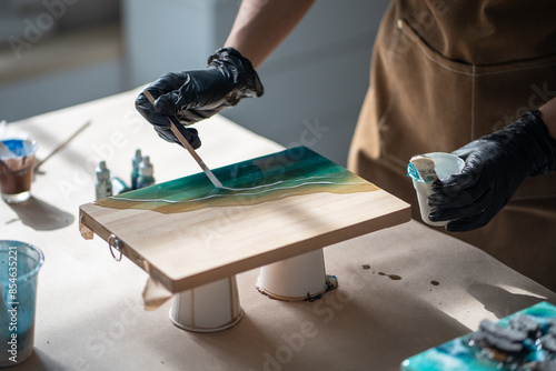 Precise neat female artist applies white pigment with stick, creates pattern of waves on epoxy resin, works on painting in technique of liquid fluid art. Woman depicts ocean, sea in creative work. photo