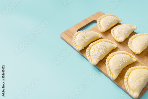 Raw samosas with sesame on a wooden on board on blue table ready for baking. Traditional Central Asian cuisine, frozen semi-finished products, copy space.