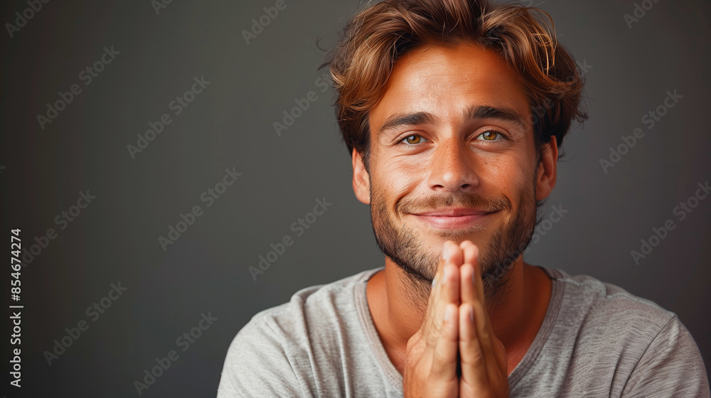 A young man with a gentle smile and brown hair stands against a gray backdrop, his hands clasped in prayer. His hopeful expression suggests a moment of deep contemplation and faith A young man with a gentle smile and brown hair stands against a gray backdrop, his hands clasped in prayer. His hopeful expression suggests a moment of deep contemplation and faith