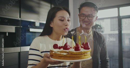 Warm moment. Asian couple celebrating birthday together. Woman blowing out candles on her birthday cake. Man hugging girl from behind. Preparing for memorable evening. Celebration. photo