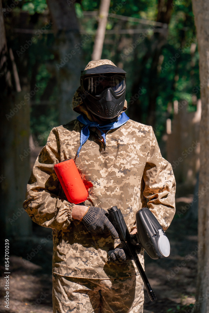Fototapeta premium in the wild near tree structures player in a pintball uniform holds a pistol with a red balloon team game camouflage clothing