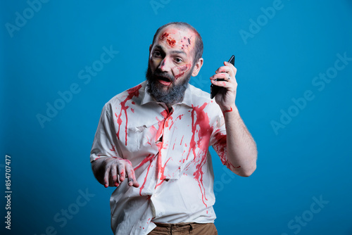 Portrait of brainless zombie holding mobile phone to ear, unable to understand technology, studio background. Mindless undead monster covered in blood and bruises listening to audio from cellphone photo