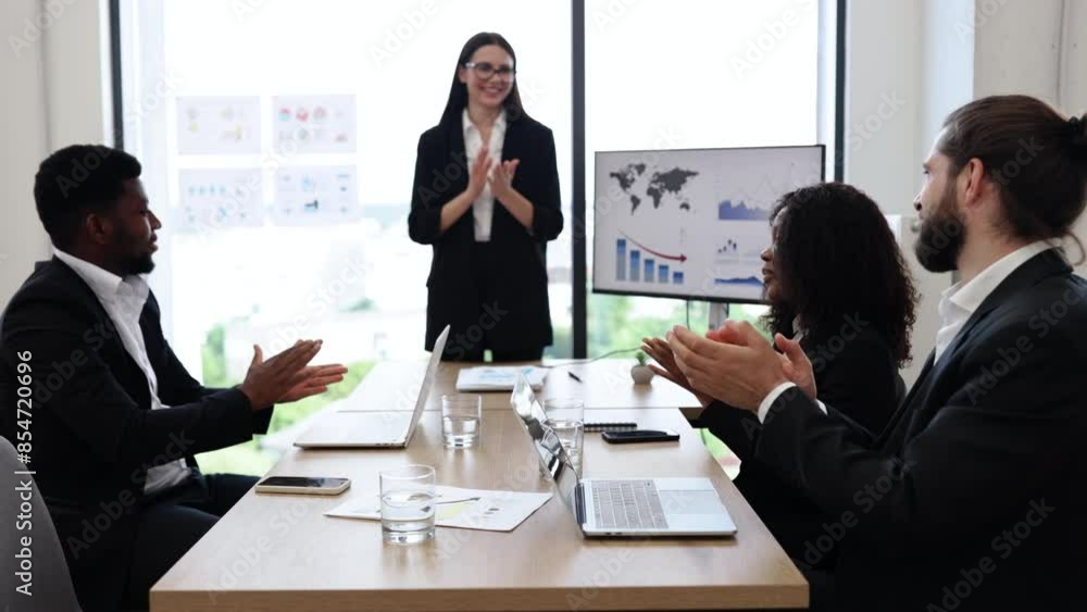 Caucasian business woman makes a report to multiracial, multinational, multicultural colleagues standing near monitor with graphs, charts. Diverse business people sit at table and clap their hands. Caucasian business woman makes a report to multiracial, multinational, multicultural colleagues standing near monitor with graphs, charts. Diverse business people sit at table and clap their hands.